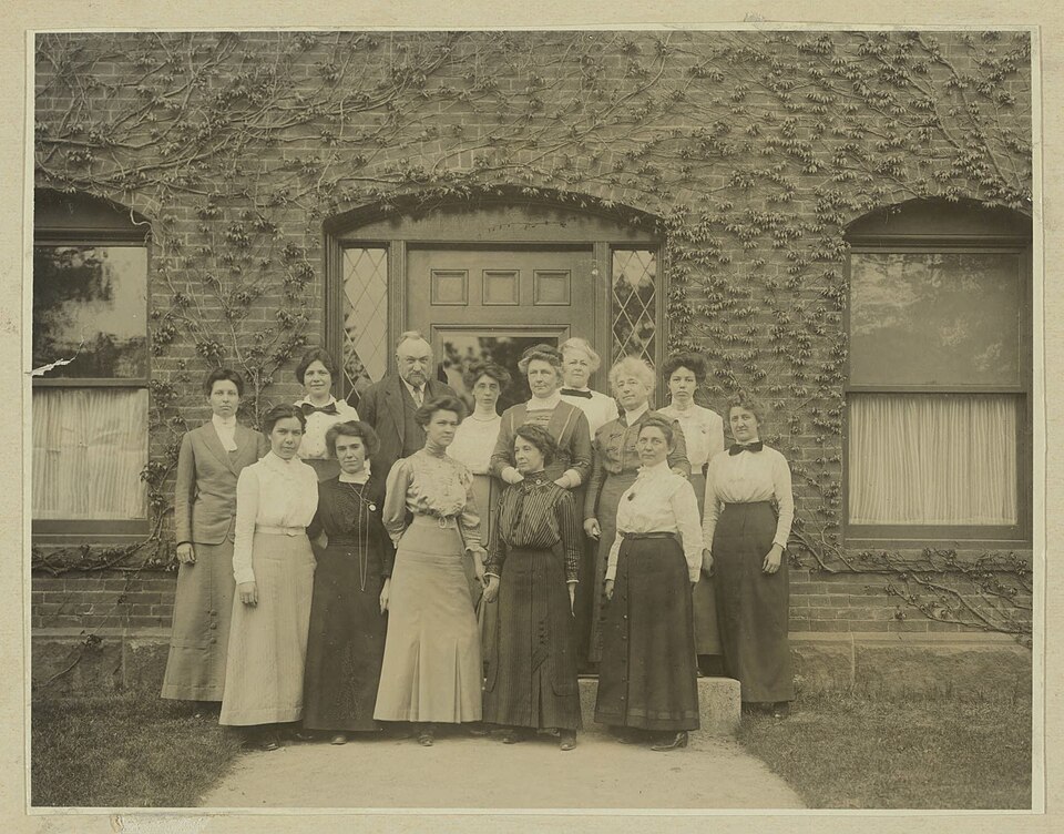 Black and white photograph taken in May 1913 showing a room of women at desks examining photographic plates and ledgers, with Edward Pickering standing at the back wearing a dark suit and a beard. A woman stands at the centre of the room, one hand raised, directing the work — most likely Annie Jump Cannon, Fleming's successor as Curator of Astronomical Photographs.