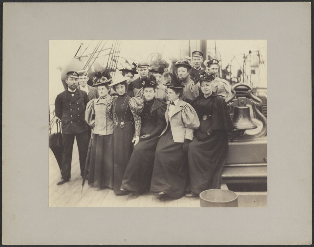 Black and white photograph from approximately 1900 showing a group of about ten women in late-Victorian dress, alongside several men in officers' uniforms, posed on the deck of a ship. A ship's bell is visible to the right.